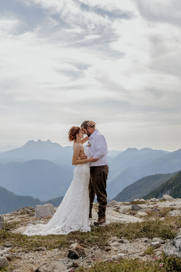 Bride and groom share a romantic kiss on a mountain peak with breathtaking views in the background.